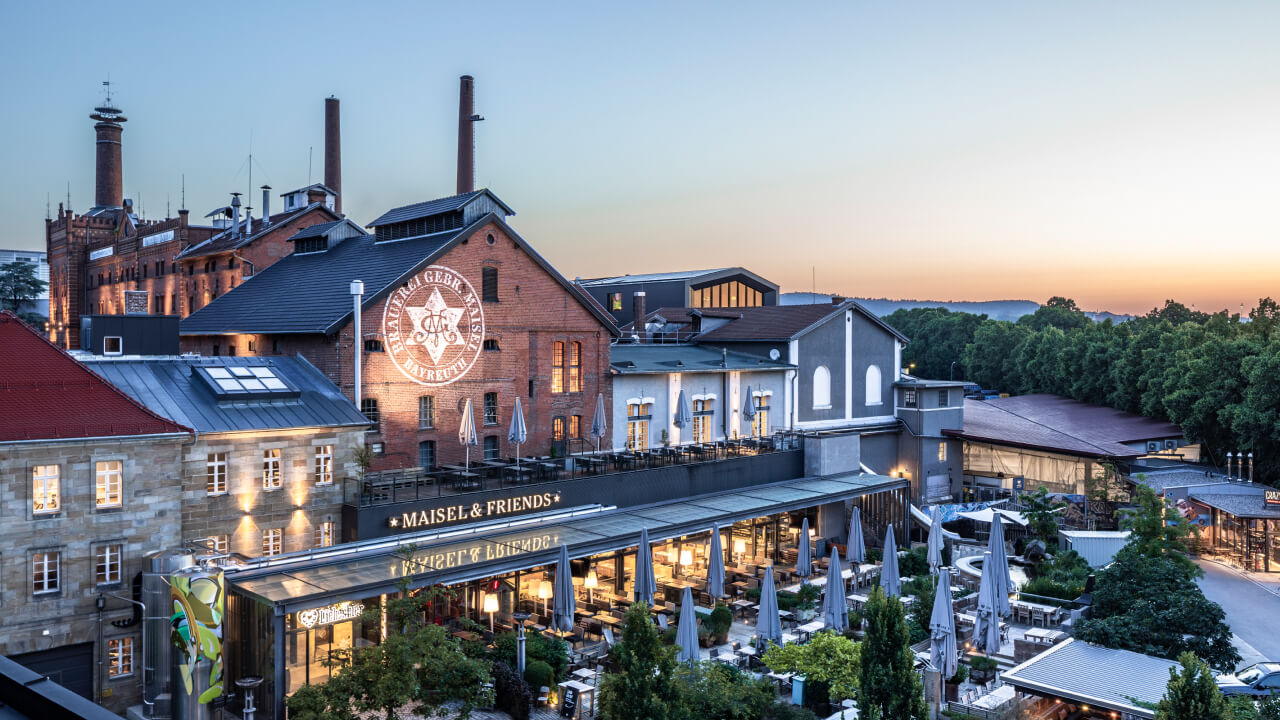 Eine historische Brauerei mit Außenbereich und Terrassen unter Sonnenschirmen, bei Dämmerung beleuchtet.