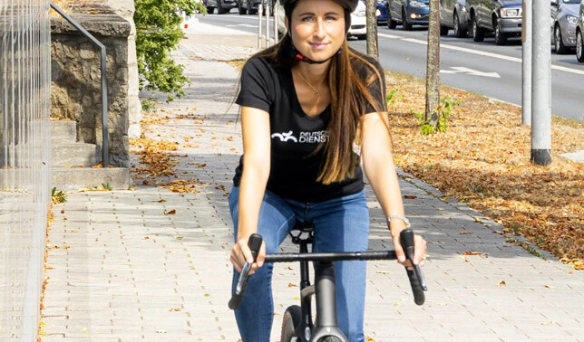 Eine Frau fährt auf einem Fahrrad auf einem Gehweg. Sie trägt einen Fahrradhelm und ein schwarzes T-Shirt mit dem Aufdruck "Deutsche Dienstrad". Im Hintergrund sind eine Straße und parkende Autos zu sehen, während auf dem Boden einige herbstliche Blätter liegen.