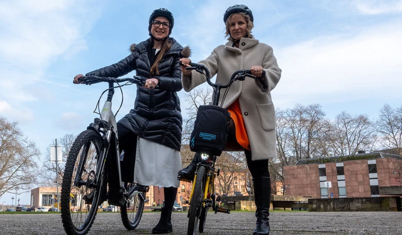 Zwei Frauen mit Helmen stehen nebeneinander und halten ihre Fahrräder. Sie lächeln in die Kamera, während sie sich im Freien befinden, mit Gebäuden und Bäumen im Hintergrund.