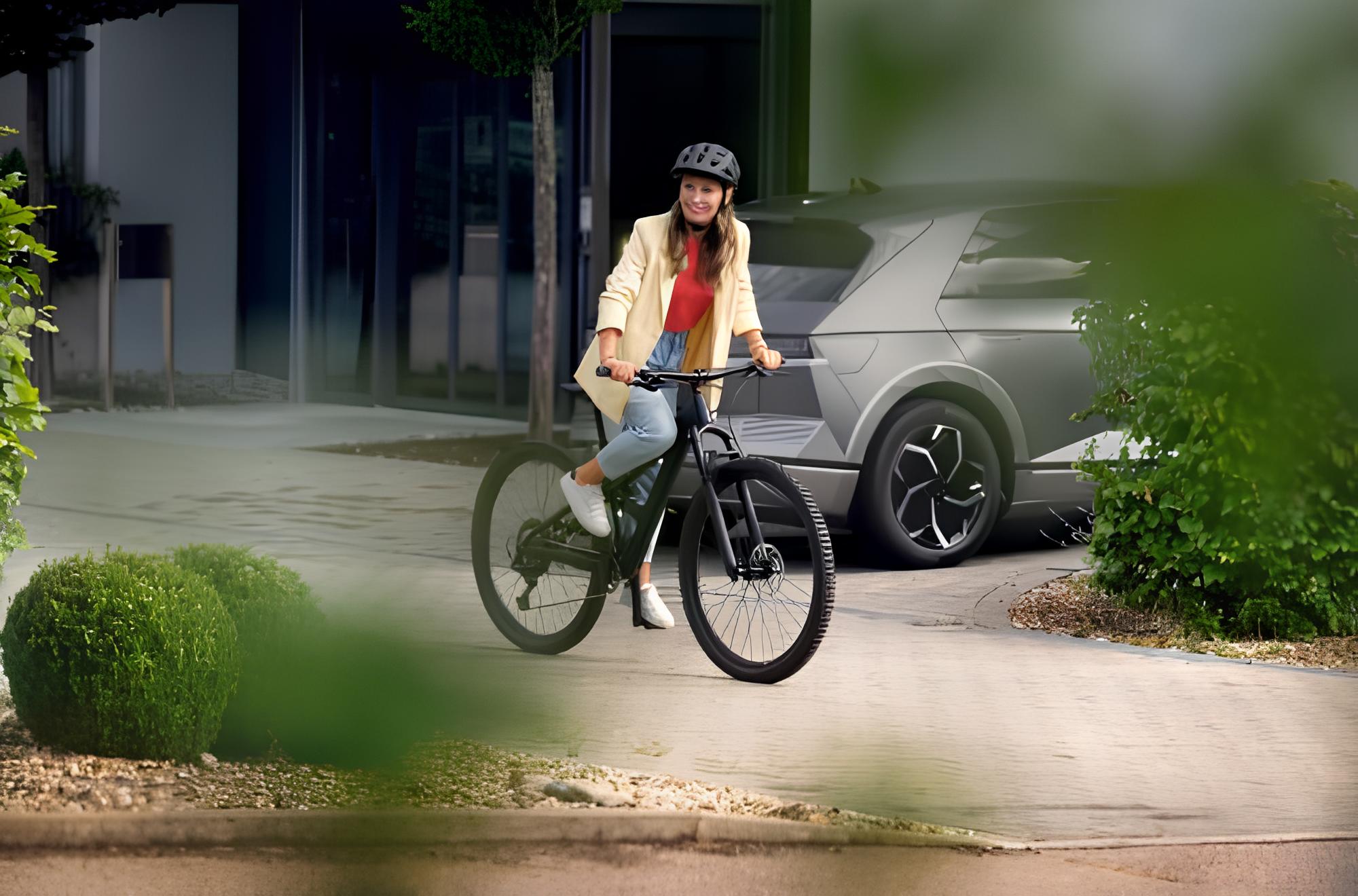 Eine Frau mit Fahrradhelm lächelt, während sie auf einem Fahrrad vor einem modernen Gebäude und einem geparkten Auto steht, umgeben von grüner Bepflanzung.