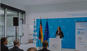 Eine Frau hält eine Rede an einem Rednerpult in einem Konferenzraum, hinter ihr stehen die Flaggen Bayerns, Deutschlands und der EU. Die Wand im Hintergrund trägt den Schriftzug „Wir bewegen Bayern“.