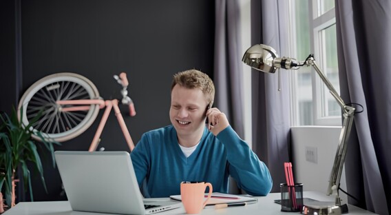 Ein Mann sitzt lächelnd an einem Schreibtisch mit Laptop, Tasse und Lampe, während ein Fahrrad im Hintergrund an der Wand lehnt.