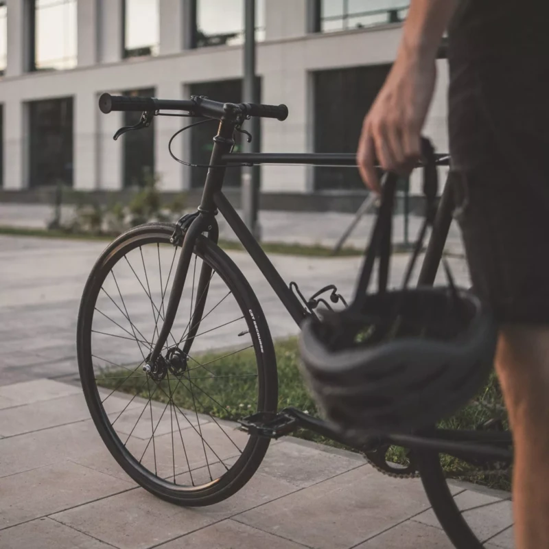 Ein stilvolles, schwarzes Fahrrad steht im Vordergrund auf einem Gehweg. Eine Person im rechten Bildbereich hält einen Fahrradhelm in der Hand, während sie neben dem Fahrrad steht. Im Hintergrund ist ein modernes Gebäude mit großen Fenstern zu sehen, das die urbane Umgebung unterstreicht.