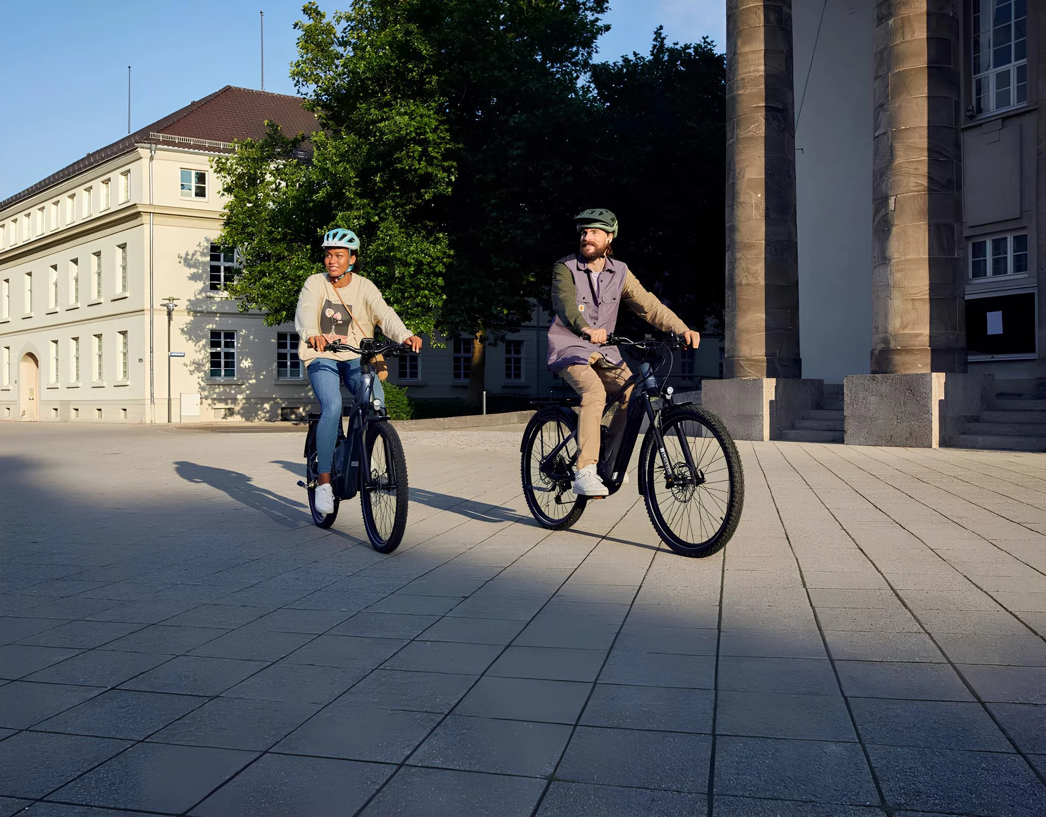 Zwei Personen fahren bei sonnigem Wetter E-Bikes auf einem gepflasterten Platz vor einem historischen Gebäude.