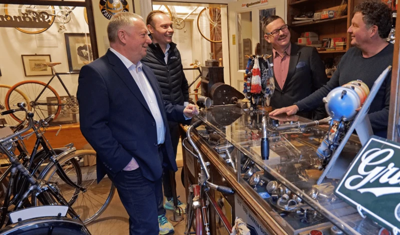 Vier Männer unterhalten sich in einem Geschäft, das mit Fahrrädern und Vintage-Gegenständen ausgestattet ist. Sie stehen um eine Glasvitrine, in der verschiedene Fahrradteile ausgestellt sind, und wirken entspannt und fröhlich.