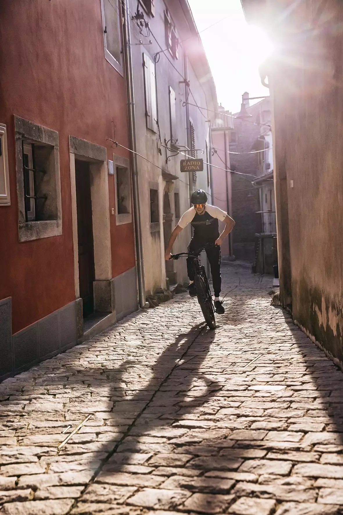 Ein Mann fährt ein E-Bike durch eine schmale, gepflasterte Gasse mit hohen Gebäuden auf beiden Seiten. Die tiefstehende Sonne wirft lange Schatten auf das Kopfsteinpflaster.