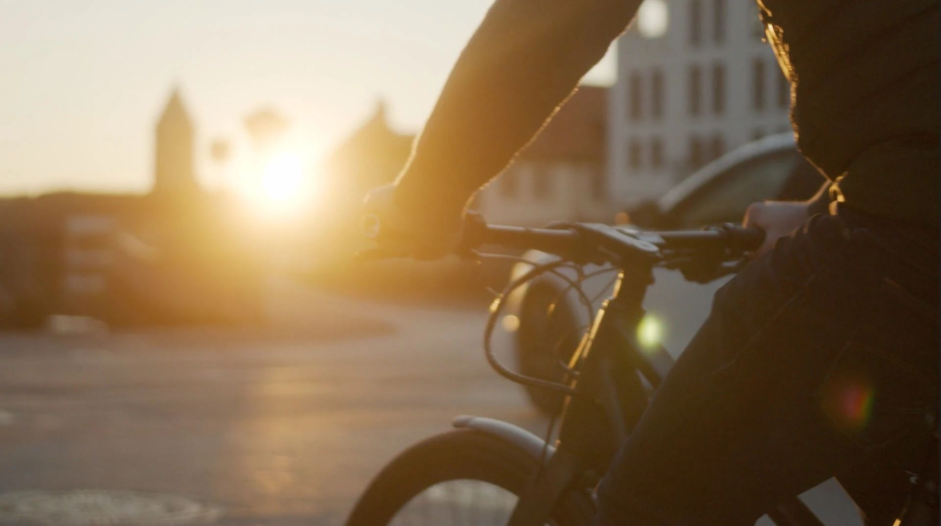 Nahaufnahme einer Person, die ein Fahrrad fährt, mit Fokus auf den Lenker. Die Szene ist bei Sonnenuntergang aufgenommen, das Licht der tiefstehenden Sonne sorgt für einen warmen und stimmungsvollen Hintergrund. Die Umgebung zeigt unscharfe Gebäude und Autos im Hintergrund.