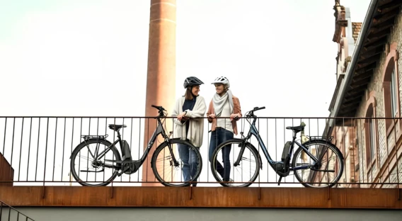 Zwei Frauen mit Fahrradhelmen lehnen an einem Geländer, neben ihren Fahrrädern, mit einem Backstein-Gebäude und Schornstein im Hintergrund.