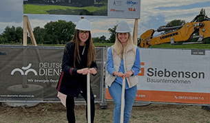 Zwei Frauen mit weißen Bauhelmen stehen auf einer Baustelle und halten Spaten in den Händen, bereit für den symbolischen Spatenstich. Hinter ihnen sind Bauzaunbanner mit Firmenlogos und ein Bagger zu sehen.