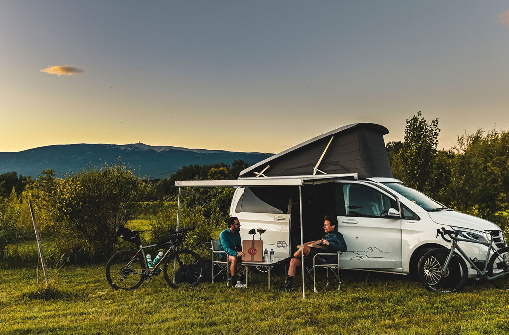 Zwei Personen sitzen vor einem weißen Camper-Van auf Campingstühlen und unterhalten sich, während ihre Fahrräder neben dem Van stehen. Im Hintergrund sind Bäume und Berge zu sehen, die Abenddämmerung sorgt für stimmungsvolle Beleuchtung.