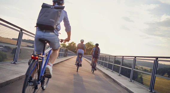 Drei Personen fahren bei Sonnenuntergang auf einem Radweg über eine Brücke. Alle tragen Helme und Rucksäcke, während sie entspannt nebeneinander herfahren.