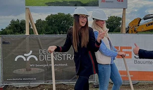 Zwei Frauen tragen Bauhelme und stehen lächelnd auf einer Baustelle mit Schaufeln in der Hand, bereit für einen symbolischen Spatenstich. Im Hintergrund sind Banner mit Logos von „Deutsche Dienstrad“ und einer Baufirma zu sehen.