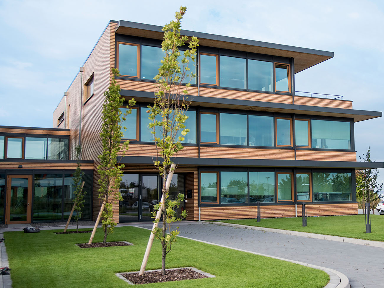 Ein modernes, dreistöckiges Gebäude mit großen Glasfenstern und einer Fassade aus Holz. Vor dem Gebäude befinden sich gepflegte Rasenflächen und junge Bäume, die in den Boden gepflanzt sind. Die Umgebung wirkt ruhig und ordentlich.