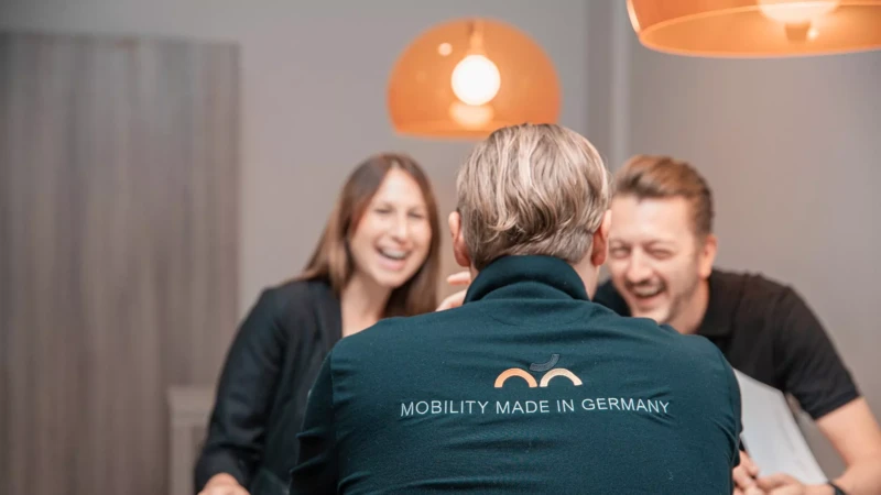 Drei Personen sitzen lachend zusammen; im Vordergrund ist der Rücken eines Mannes mit der Aufschrift 'MOBILITY MADE IN GERMANY' auf seinem Pullover zu sehen.