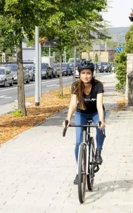 Eine Frau trägt einen Fahrradhelm und lächelt in die Kamera, während sie ein Fahrrad hält. Sie ist in einem grauen Blazer und einem grünen Pullover gekleidet und steht auf einer sonnigen Straße.