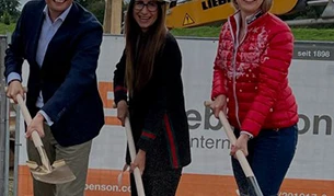 Drei Personen in Arbeitskleidung und mit weißen Bauhelmen führen auf einer Baustelle einen symbolischen Spatenstich durch. Hinter ihnen ist ein Bauzaun mit Firmenlogos sowie ein Bagger zu sehen.