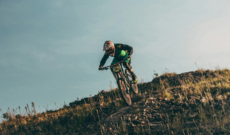 Ein Mountainbiker mit Schutzhelm fährt einen steilen, felsigen Hang hinunter. Er trägt eine grüne und schwarze Sportbekleidung. Der Himmel im Hintergrund ist klar, und die Szenerie zeigt eine trockene, grasbewachsene Landschaft.