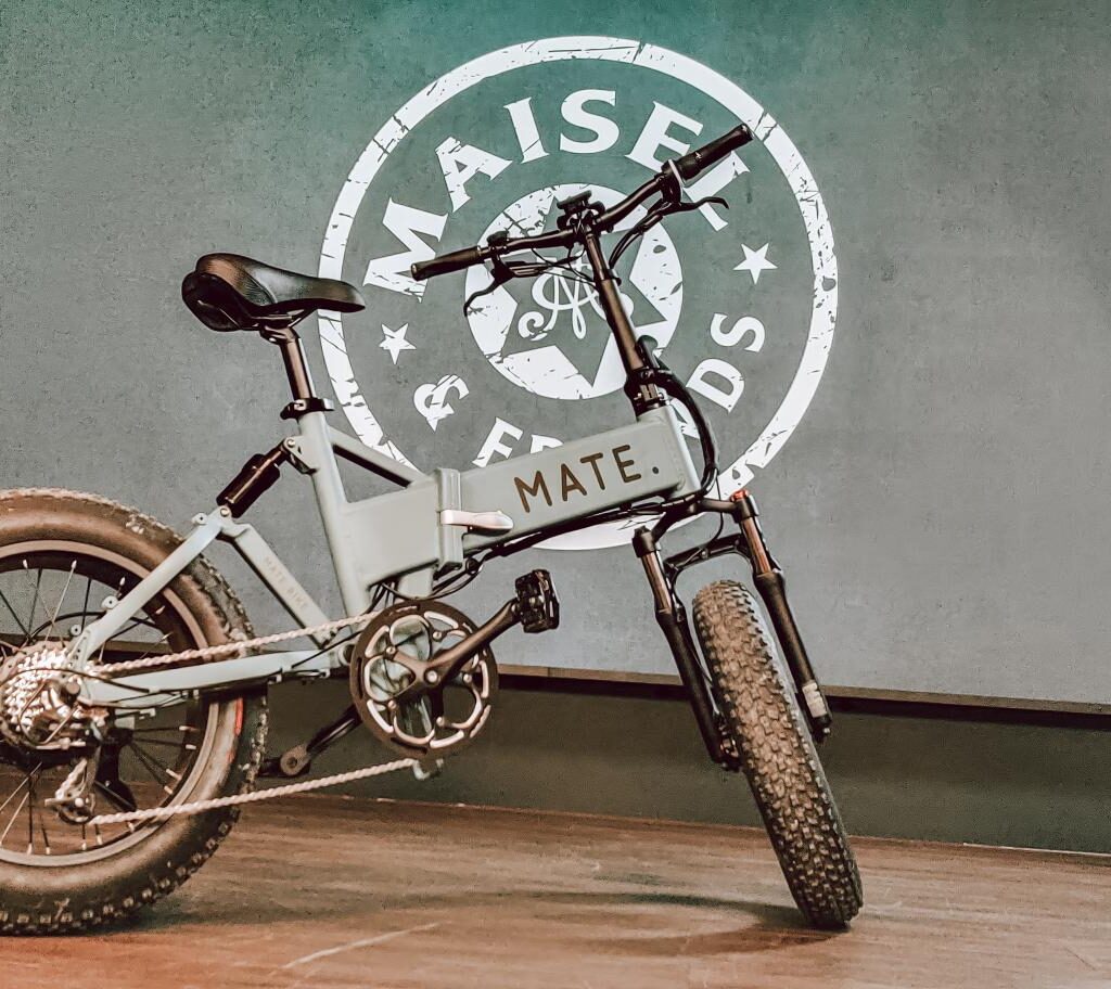 Ein graues E-Bike der Marke MATE steht vor einer Wand mit dem Logo "Maisel & Friends". Das Fahrrad hat dicke Reifen und einen stabilen Rahmen.