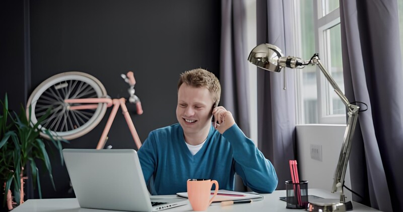 Ein Mann sitzt lächelnd an einem Schreibtisch mit Laptop, Tasse und Lampe, während ein Fahrrad im Hintergrund an der Wand lehnt.