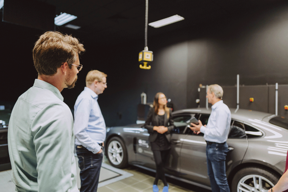 Vier Personen stehen in einem dunklen Raum, diskutieren und betrachten ein graues Auto.