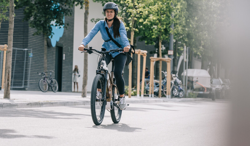 Eine Frau fährt auf einem E-Bike durch eine Straße in einer städtischen Umgebung, dabei trägt sie einen Helm und eine blaue Jacke. Im Hintergrund sind Bäume und weitere Fahrräder zu sehen.