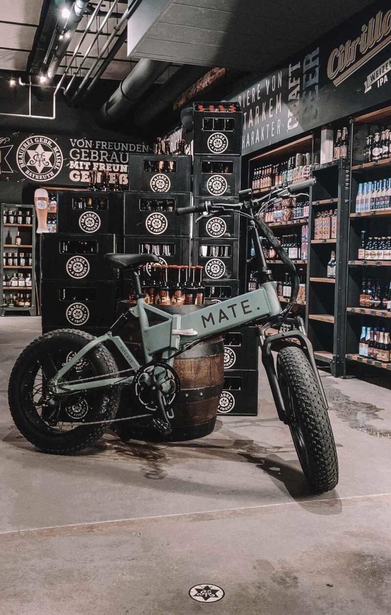 Ein graues MATE E-Bike steht vor einem Holzfass in einem Geschäft für Craft-Biere. Im Hintergrund sind Bierkisten und Regale mit verschiedenen Biersorten zu sehen.