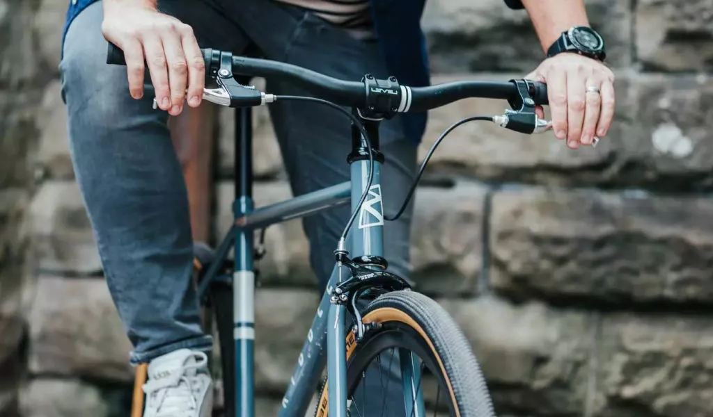 Nahaufnahme eines Fahrradlenkers mit Händen eines Radfahrers, der das Rad hält. Der Fahrer trägt graue Hosen, helle Schuhe und eine Uhr am linken Handgelenk. Im Hintergrund sind grob behauene Steinwände zu sehen.