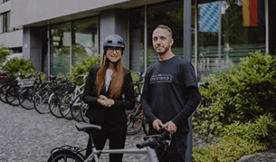 Eine Frau mit Helm und ein Mann stehen neben einem Fahrrad vor einem modernen Gebäude, im Hintergrund sind weitere Fahrräder sowie Bäume zu sehen. Die Frau trägt einen Anzug, der Mann ein dunkles T-Shirt.