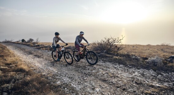 Zwei Personen fahren mit Mountainbikes auf einem Schotterweg bei Sonnenaufgang oder Sonnenuntergang in einer offenen, naturbelassenen Landschaft.