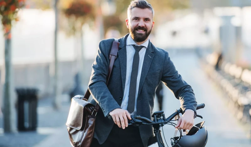 Ein lächelnder Mann in Anzug und Krawatte hält ein Fahrrad. Er trägt eine braune Umhängetasche und hält einen Fahrradhelm in der Hand. Im Hintergrund sind verschwommene Bäume und städtische Elemente zu sehen, was auf eine Szene in der Stadt hindeutet.