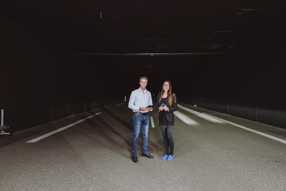 Zwei Personen stehen nebeneinander in einem dunklen, tunnelartigen Raum auf einer Straße mit Markierungen. Sie schauen in die Kamera und halten Gegenstände in den Händen.