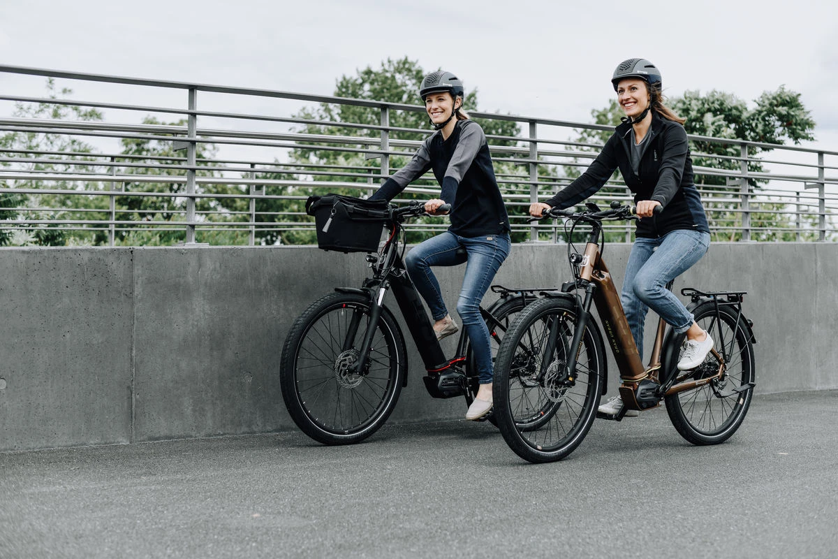 Zwei Frauen in Helmen und Freizeitkleidung fahren nebeneinander auf E-Bikes entlang einer Betonmauer mit Metallgeländer. Sie lächeln und genießen ihre Fahrt auf einem bewölkten Tag.