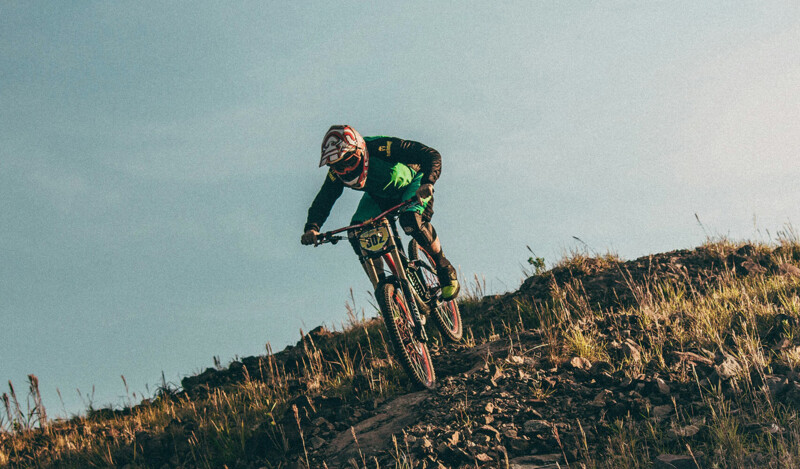 Ein Mountainbiker mit Schutzhelm fährt einen steilen, felsigen Hang hinunter. Er trägt eine grüne und schwarze Sportbekleidung. Der Himmel im Hintergrund ist klar, und die Szenerie zeigt eine trockene, grasbewachsene Landschaft.