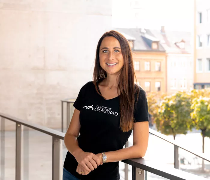 Eine Frau steht lächelnd auf einer Terrasse und blickt in die Kamera. Sie trägt ein schwarzes T-Shirt mit dem Logo von "Deutsche Dienstrad". Im Hintergrund sind Gebäude und Bäume zu sehen, die im Sonnenlicht stehen.