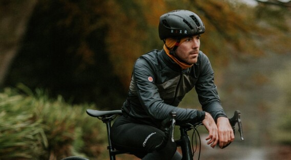 Ein Mann in kompletter Fahrradbekleidung sitzt auf einem schwarzen Rennrad und lehnt sich auf den Lenker. Er trägt einen Helm und ein Halstuch, während er auf einem feuchten, von Herbstlaub bedeckten Weg inmitten grüner Vegetation fährt.