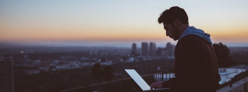 Ein Mann arbeitet an einem Laptop im Freien bei Sonnenuntergang, mit Blick auf eine Stadt im Hintergrund.