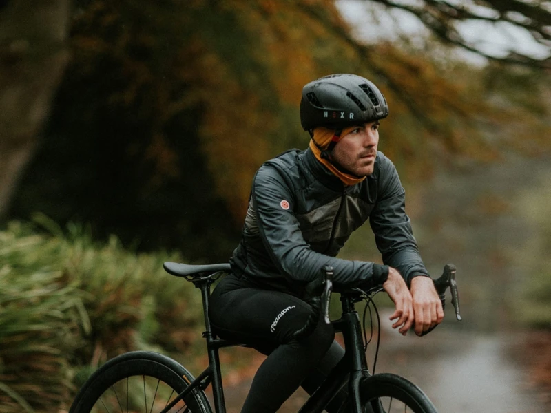 Ein Mann in kompletter Fahrradbekleidung sitzt auf einem schwarzen Rennrad und lehnt sich auf den Lenker. Er trägt einen Helm und ein Halstuch, während er auf einem feuchten, von Herbstlaub bedeckten Weg inmitten grüner Vegetation fährt.