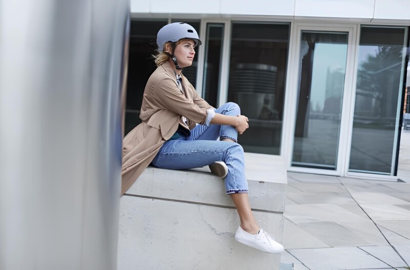 Eine Frau mit Fahrradhelm sitzt entspannt auf einer Steinmauer in einem urbanen Umfeld. Sie trägt eine helle Jeans, weiße Sneaker und einen beigen Mantel und schaut gedankenverloren zur Seite.