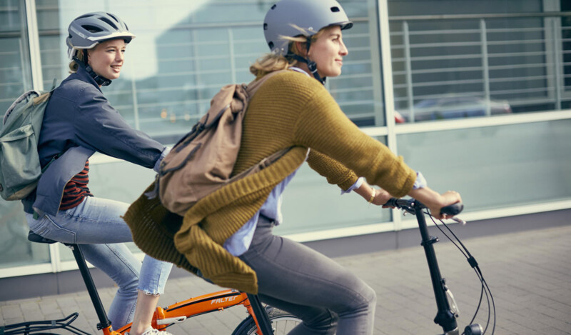 Zwei Frauen fahren auf Fahrrädern, beide tragen Helme und Rucksäcke. Die Szene spielt in einer modernen städtischen Umgebung, im Hintergrund sind Glasfassaden zu sehen.