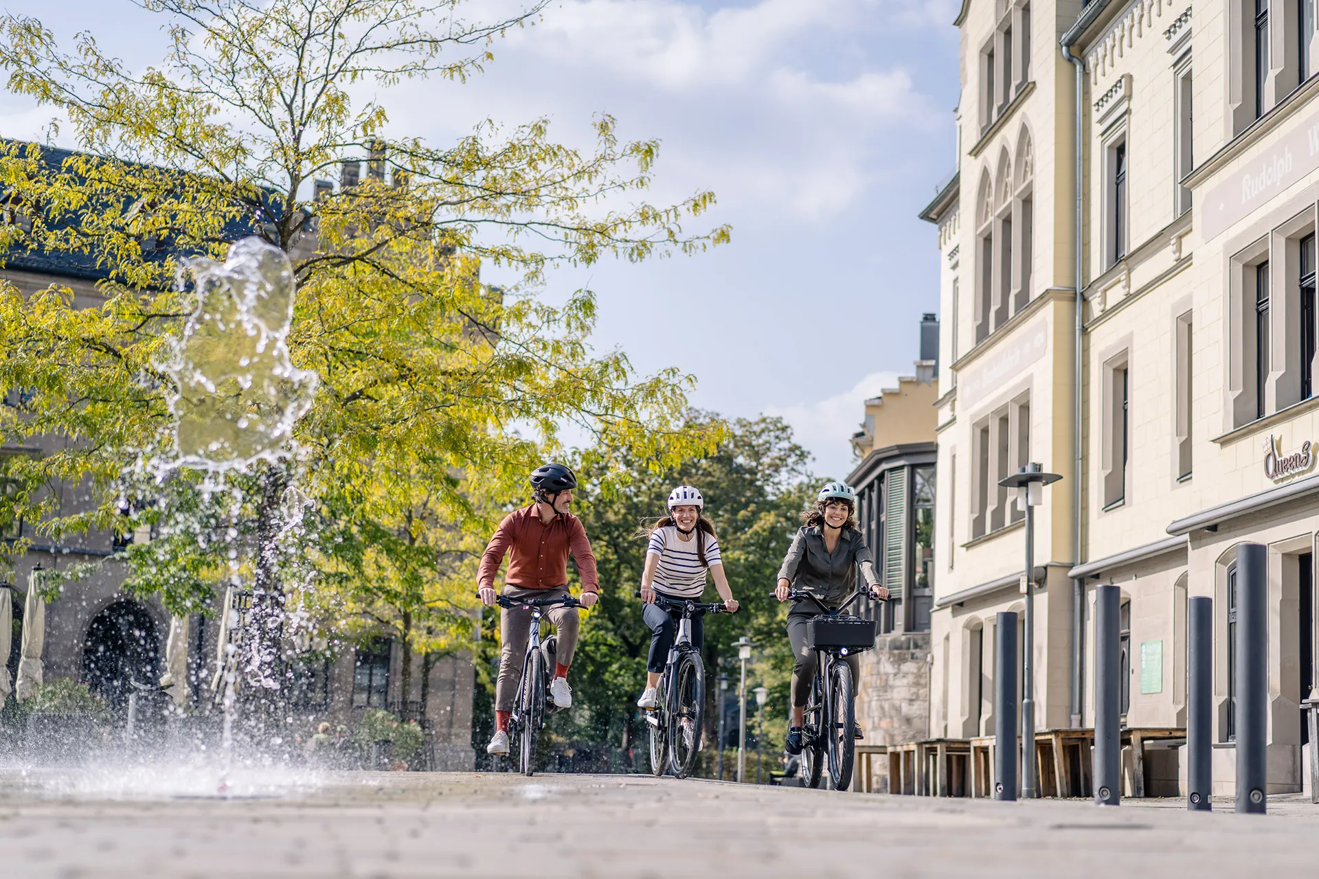 Eine Gruppe von Menschen fahren auf einem Fahrrad auf einem Gehweg entlang, und ihre Spiegelung ist auf der Glaswand rechts neben ihr zu sehen.