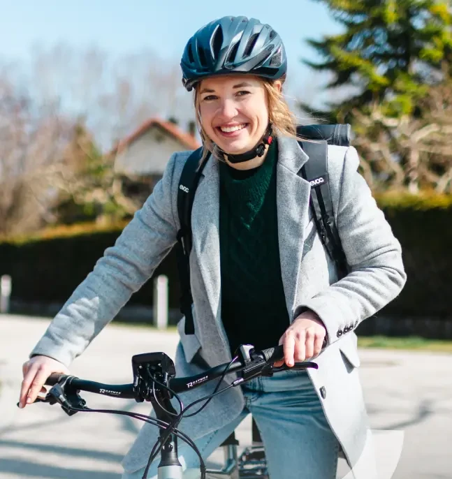 Eine Frau trägt einen Fahrradhelm und lächelt in die Kamera, während sie ein Fahrrad hält. Sie ist in einem grauen Blazer und einem grünen Pullover gekleidet und steht auf einer sonnigen Straße.