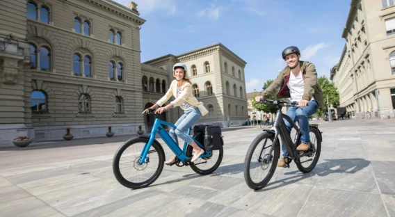 Ein Mann und eine Frau fahren lächelnd mit Fahrrädern über einen Platz. Beide tragen Helme und Freizeitkleidung. Im Hintergrund befinden sich historische Gebäude mit großen Fenstern und Steinfassaden unter einem klaren blauen Himmel.