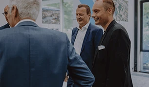 Mehrere Männer in Anzügen unterhalten sich in einem modernen Konferenzraum. Sie wirken gut gelaunt und stehen in einem lockeren Gespräch beieinander.