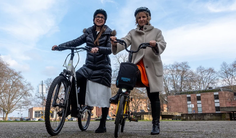 Zwei Frauen mit Helmen stehen nebeneinander und halten ihre Fahrräder. Sie lächeln in die Kamera, während sie sich im Freien befinden, mit Gebäuden und Bäumen im Hintergrund.