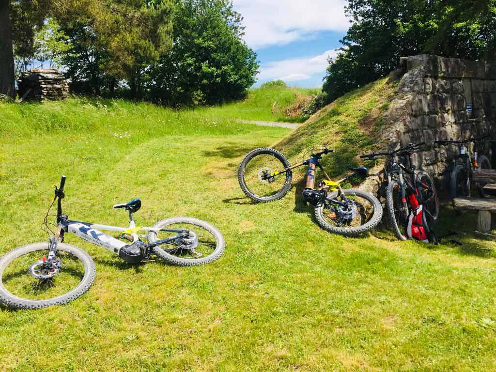  Mehrere Fahrräder, darunter Mountainbikes, liegen und stehen auf einer grasbewachsenen Fläche im Freien, umgeben von Bäumen und einem alten Steinbau. Sonniges Wetter mit klarem Himmel im Hintergrund.