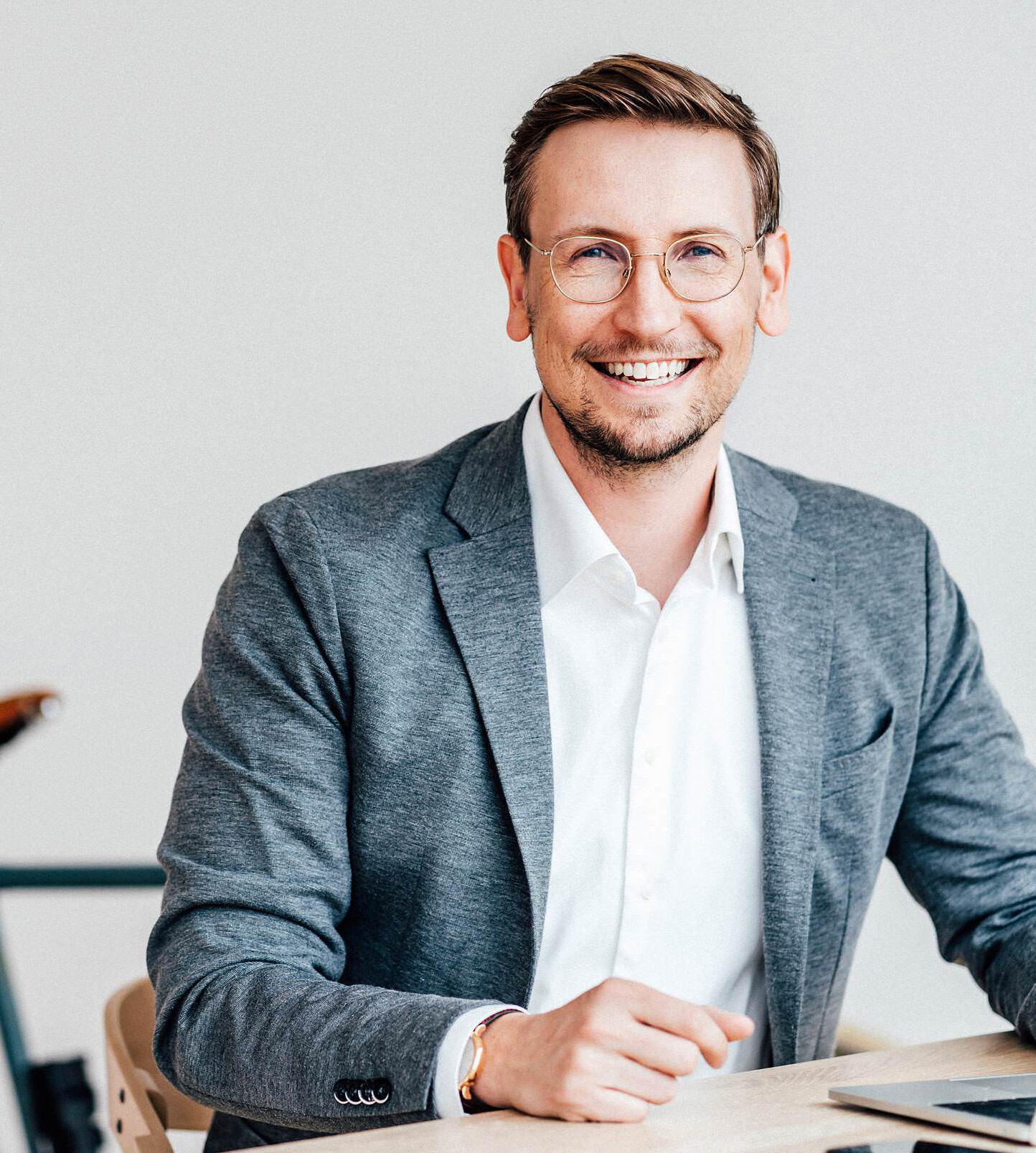 Ein lächelnder Mann mit Brille und hellem Hemd sitzt an einem Tisch. Er trägt ein graues Sakko und schaut direkt in die Kamera.