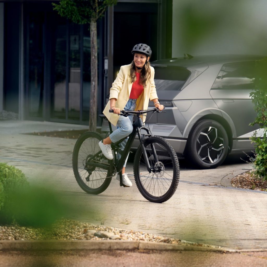 Eine Frau mit Helm sitzt lächelnd auf einem Fahrrad, umgeben von einem modernen städtischen Umfeld mit Pflanzen im Vordergrund.