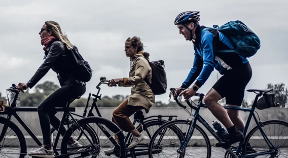 Drei Personen fahren auf Fahrrädern in einer Reihe: Eine Frau mit Schal und Lederjacke, eine Frau in beigefarbenem Mantel, und ein Mann in sportlicher Fahrradkleidung mit Helm. Alle haben Rucksäcke und fahren auf einem asphaltierten Weg an einem bewölkten Tag.