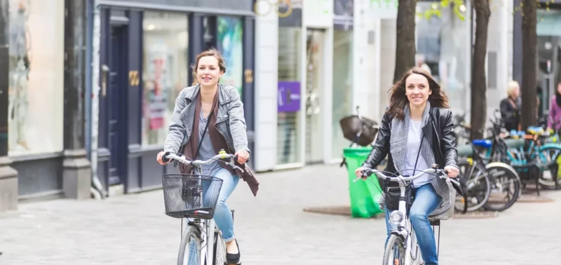 Zwei Frauen fahren lächelnd mit Fahrrädern durch eine Fußgängerzone.