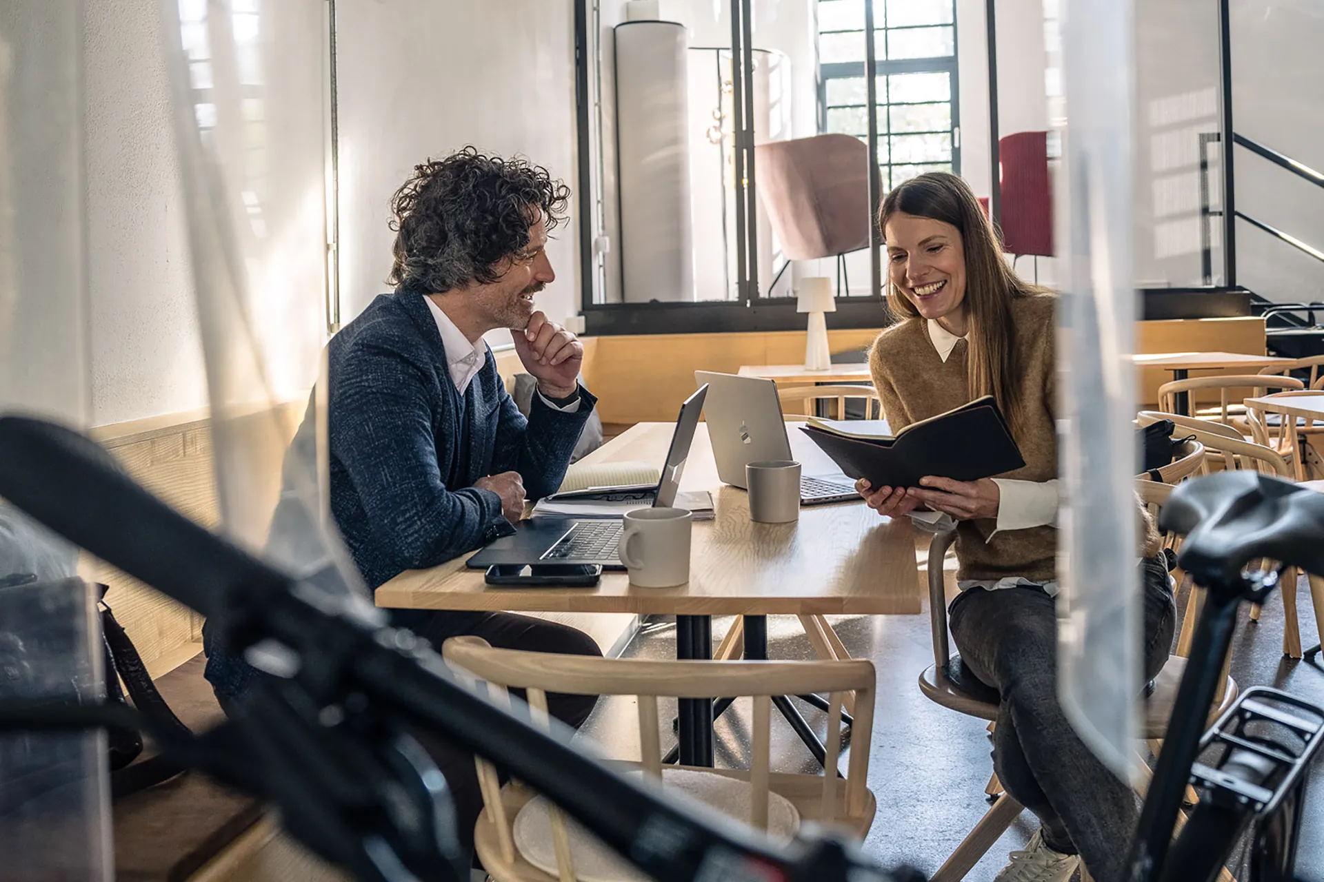 Eine Frau und ein Mann sitzen an einem Tisch, zwei Laptops vor ihnen und besprechen offene Punkte.